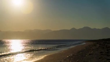 Zaman ayarlı, denizde gün batımı. Güneş dağların arkasında batıyor. Kumlu boş bir plajda.