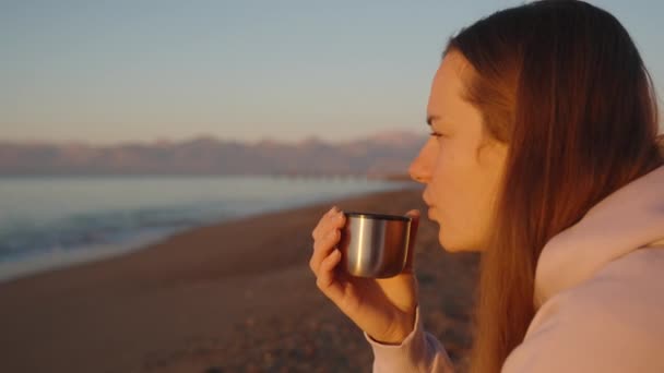 海上清早 一个年轻的女人喝热饮从一个热水瓶 在高山的背景下日出 — 图库视频影像