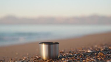 Denizde soğuk bir sabah, termostan bir kupa sahildeki küçük taşların üzerinde duruyor. Yakın plan..