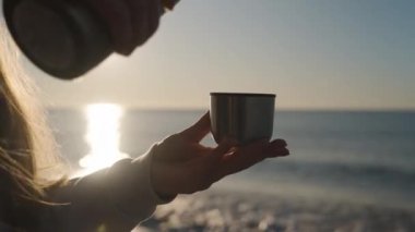 Denizde gün doğumunda genç bir kadın sıcak çayı termostan metal bardağa dolduruyor. Güneş ve dalgaların arkaplanına karşı.