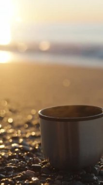Sıcak içeceği olan bir termostan bir kupa kayalıklarda, deniz dalgalarının yanında duruyor. Soğuk bir sabah, Gündoğumu. Kameranın odak noktası bardaktan güneşe. Dikey video Sosyal Ortamı