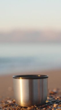 Denizde soğuk bir sabah, termostan bir kupa sahildeki küçük taşların üzerinde duruyor. Yakın plan. Dikey video Sosyal Ortamı