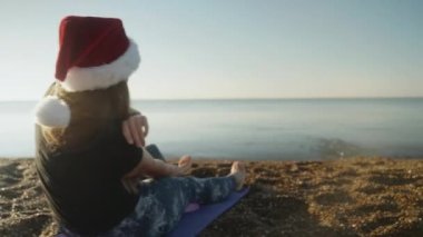 Kırmızı Noel Baba şapkalı genç bir kadın kumlu sahilde oturuyor ve taş atıyor..
