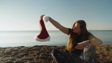 Genç bir kadın sabahın erken saatlerinde sahilde oturur ve elinde kırmızı Noel Baba şapkasını sallar..