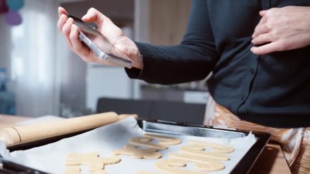 女性は異なる形のさまざまなジンジャーブレッドクッキーでベーキングシートの写真を撮るために彼女の携帯電話を使用しています — ストック動画