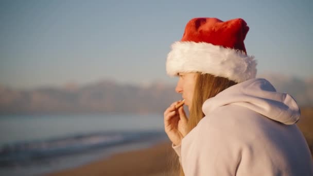 一个年轻的女人在日出时坐在海边 享受着圣诞姜饼饼干和茶 背景是群山 — 图库视频影像