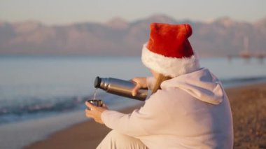 Dağların ve denizin önünde sabahın erken saatlerinde Noel Baba şapkalı genç bir kadın termostan çay döker..