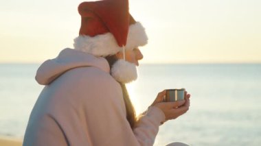 Deniz kenarında serin bir Noel sabahı, kırmızı Noel Baba şapkalı genç bir kadın sıcak çay içiyor. Kadehten buhar yükseliyor..