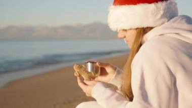 Noel Baba şapkalı genç bir kadın, zencefilli kurabiyelere erkek şeklinde bakar ve elinde bir kupa çay ile şafak vakti deniz kenarında oturur..