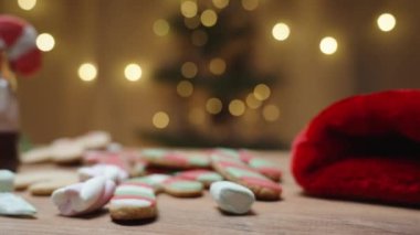 Marşmelovlu zencefilli kurabiye ve bir fincan sıcak çikolata masaya saçıldı. Arka planda çelenkleri olan bir Noel ağacı var. Sıcak ve rahat bir ev ortamı yaratıyor..