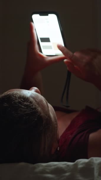 若い女性はベッドの端に横たわり 携帯電話垂直 ソーシャルメディアを保持しています — ストック動画