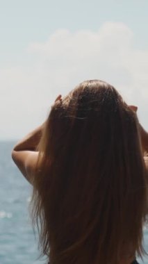 Deniz kenarındaki mayo giymiş tombul genç kadın topuz şeklinde uzun saçlar topluyor. Dikey, Sosyal Ortam.