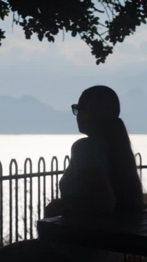 Genç kadın bir ağacın altında bir masada oturuyor, denizi ve dağları izliyor. Dikey, Sosyal Ortam.