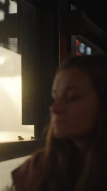 Deniz kenarındaki bir kafede pencereden dışarı bakan genç bir kadın. Arka planda, gün batımı ve suyun üzerindeki güneşten gelen ışık. Dikey video Sosyal Ortamı.