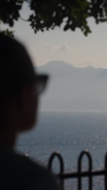 一个年轻的女人 戴着眼镜站在树荫下 望着大海和群山 社交媒体 — 图库视频影像