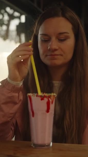 カフェにいる若い女性は イチゴのミルクシェイクを 雲の中を太陽の光線の後ろに わらを通して飲んでいる 垂直ビデオ ソーシャルメディア — ストック動画