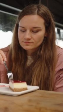Genç bir kadın bir kafede ceketle oturmuş peynirli kek yiyor. Masada çilekli milkshake var. Dikey video Sosyal Ortamı.
