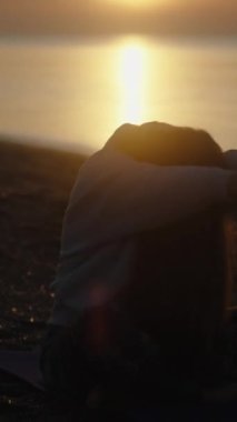 Şafak vakti deniz kıyısında yoga. Genç kadın halıya oturuyor ve ellerini güneşe uzatıyor. Dikey video Sosyal Ortamı.