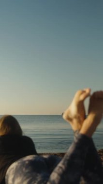 Şafakta deniz. Balıkçı teknesi ve sahilde ayakları havada yatan genç bir kadın. Dikey video Sosyal Ortamı.