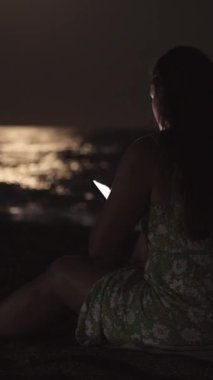 Elbise giymiş genç bir kadın kumsalda deniz kenarında oturur ve akıllı telefona bakar. Dikey, Sosyal Ortam