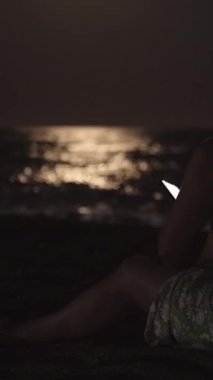 Elbise giymiş genç bir kadın kumsalda deniz kenarında oturur ve akıllı telefona bakar. Dikey, Sosyal Ortam