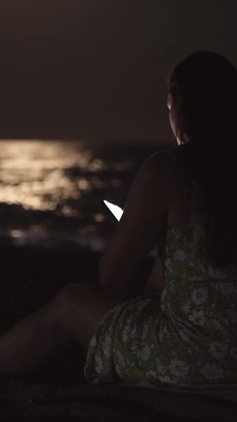 一位身穿连衣裙的年轻女子坐在海边的海边 凝视着智能手机 社交媒体 — 图库视频影像