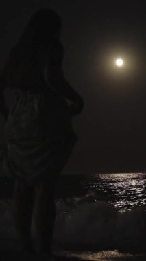 Genç kadın gece plajda duruyor ve ayaklarını suya sokuyor. Dikey, Sosyal Ortam