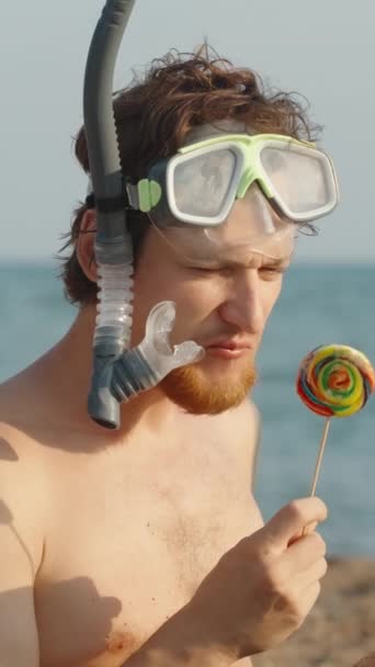 一个男人带着红胡子 戴着水肺护目镜 拿着烟斗 在海滩上舔着一个五颜六色的棒棒糖 看着它 — 图库视频影像