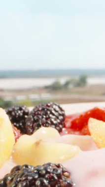 Güneşli bir sabahta balkonda açık gökyüzünün altında kahvaltı. Meyve ve yoğurt tabağı. Kaşıkla topluyorum. Dikey. Sosyal Medya