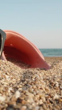 Güneş gözlüğü ve pembe parmak arası terlik denizin arka planında kumda yatıyor. Güneş gözlüklere yansıyor. Dikey. Sosyal Medya