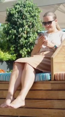 Genç kadın büyük bir şemsiyenin altında ahşap bir bankta oturuyor. Telefonuna bakar ve gülümser. Dikey. Sosyal Medya