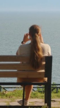 Genç kadın sahildeki bir bankta oturuyor ve manzaranın fotoğraflarını çekiyor. Dikey. Sosyal Medya