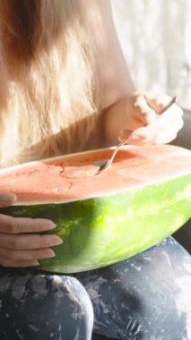 Genç kadın dizlerinin üzerinde büyük bir karpuz tutuyor ve hamuru kaşıkla yiyor. Dikey. Sosyal Medya