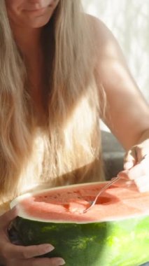 Genç kadın dizlerinin üzerinde büyük bir karpuz tutuyor ve hamuru kaşıkla yiyor. Dikey. Sosyal Medya