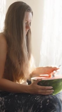 Genç kadın büyük bir karpuzla kanepede oturur, kaşıkla yer. Dikey. Sosyal Medya