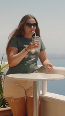 Güneş gözlüklü genç bir kadın buzlu kahve içer. Arkasında deniz var. Dikey. Sosyal Medya