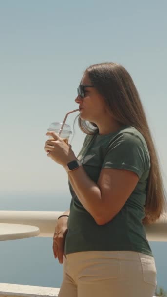 サングラスを持った若い女性がレールに立ってアイスコーヒーを飲む その背後には海がある 垂直に ソーシャルメディア — ストック動画