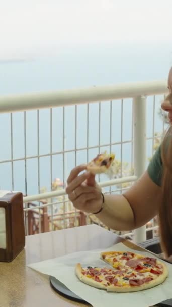 山の上のカフェに座っている若い女性 海の美しい景色を眺めることができます 彼女はピザを食べている ソーシャルメディア — ストック動画