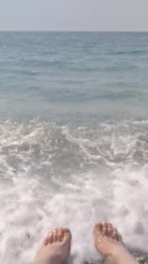 Deniz kıyısında genç bir kadının ayakları, bir dalga. Dikey video.