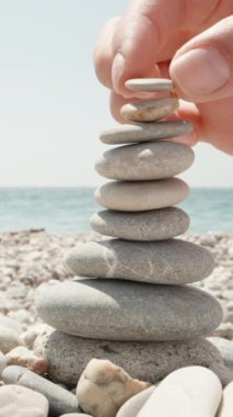 Yakın plan, bir kadın cairn yapıyor. Deniz kıyısında el yapımı taş kule. Sahilde bir yığın dengeli çakıl taşı. Denizin arka planına karşı Dikey video.