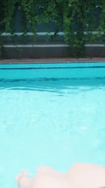 Mavi havuz, güneşli bir gün. Genç bir kadın ayaklarını suya sokuyor. Dikey video..