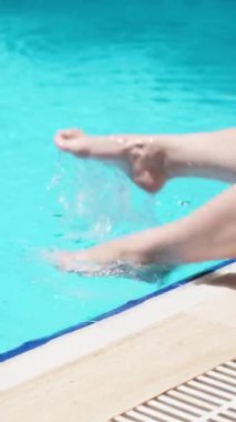 Mavi havuz, güneşli bir gün. Genç bir kadın ayaklarını suya sokuyor. Dikey video..