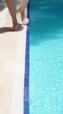 Mavi havuz, güneşli bir gün. Genç bir kadın havuz kenarında ayağını suya daldırıyor. Dikey video..