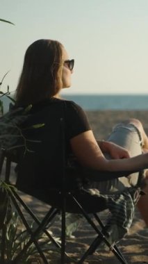 Sahilde elinde bir bardak şarapla gün batımını izleyen genç bir kadın. Piknik, kumda barbekü. Dikey video Sosyal Ortamı
