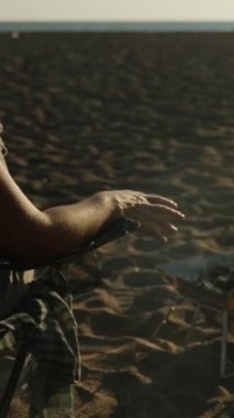 Deniz kenarındaki sahilde genç bir kadın şöminenin yanındaki sandalyede oturuyor, gün batımını seyrediyor ve kendini ateşin yanında ısıtıyor. Dikey video Sosyal Ortamı