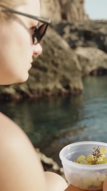 一个穿着泳衣的年轻女子坐在岩石上吃葡萄 她把手指指向大海 垂直视频社交媒体 — 图库视频影像