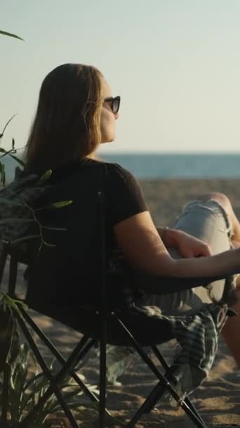 一位年轻女子在海边的海边 手里拿着一杯葡萄酒 看着夕阳西下 在沙滩上烧烤 垂直视频社交媒体 — 图库视频影像