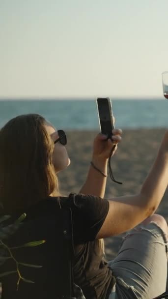 一位在海边的年轻女子在夕阳西下拍下了手中拿着一杯葡萄酒的照片 海滩上的野餐 垂直视频社交媒体 — 图库视频影像