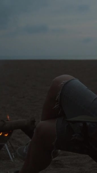 在海滩上野餐 黄昏和篝火熊熊燃烧 一位坐在椅子上的年轻女子拿着一杯葡萄酒和棉花糖放在手杖上 垂直视频社交媒体 — 图库视频影像
