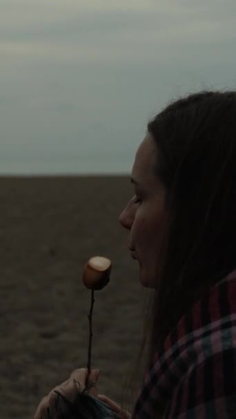海岸で夕方のピクニックを手配する 若い女性はフライドマシュマロ 垂直ビデオソーシャルメディアのために吹き 食べます — ストック動画